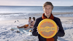 Man holding a Jarlsberg cheese wheel while a couple sits on the beach in the background