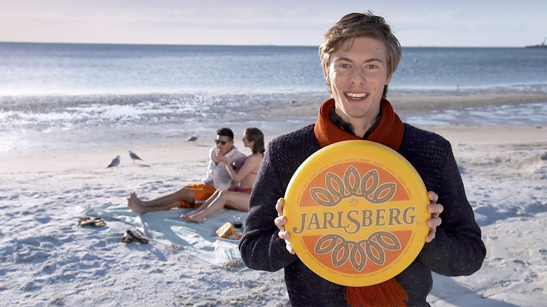 Man holding a Jarlsberg cheese wheel while a couple sits on the beach in the background