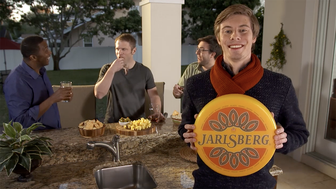 Man holding a Jarlsberg cheese wheel while people enjoy snacks in the background