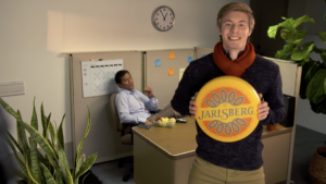 Man holding a Jarlsberg cheese wheel while a man snacks in the office in the background