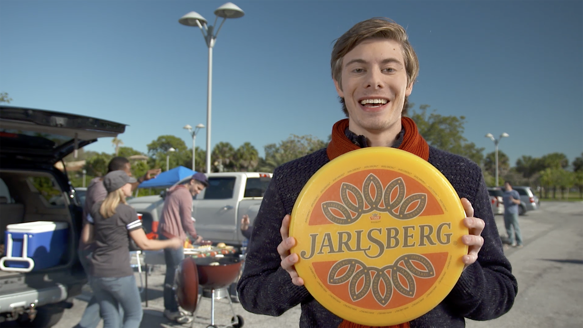 Man holding a Jarlsberg cheese wheel while people tailgate in the background