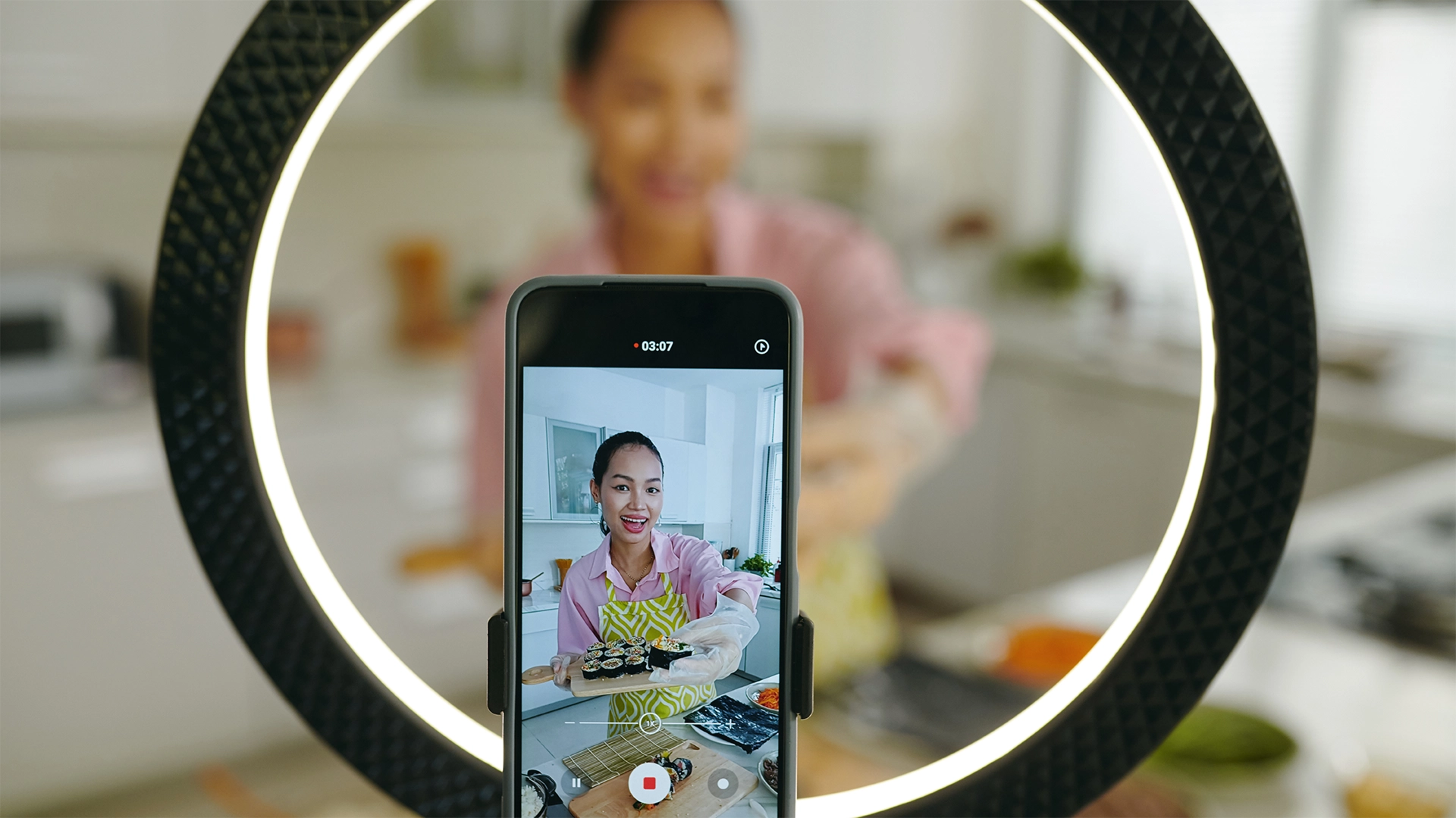 A woman filming a cooking video on her smartphone, smiling and holding a tray of sushi, with a ring light in the foreground.