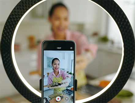 A woman filming a cooking video on her smartphone, smiling and holding a tray of sushi, with a ring light in the foreground.