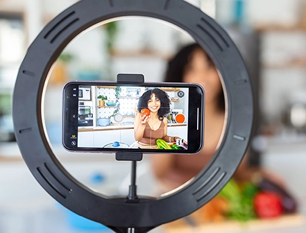 Woman holding fruit in a kitchen, recording a video on her phone with a ring light, showing how personable nano-influencers are.