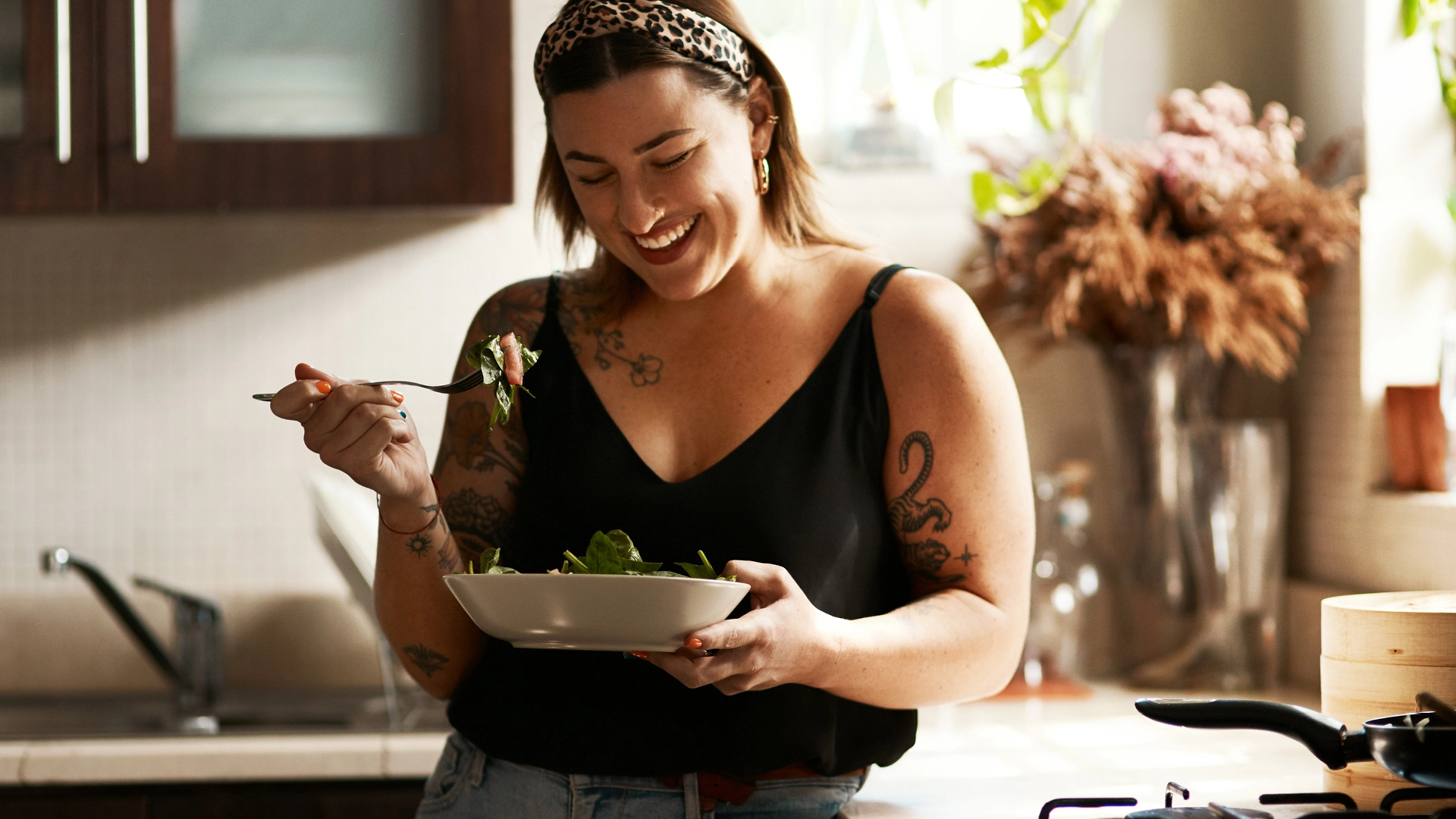 A woman smiling & laughing while getting ready to enjoy her bowl of salad in a kitchen, highlighting the happiness and joy she gets from eating healthy and focusing on her gut health.