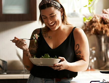 A woman smiling & laughing while getting ready to enjoy her bowl of salad in a kitchen, highlighting the happiness and joy she gets from eating healthy and focusing on her gut health.